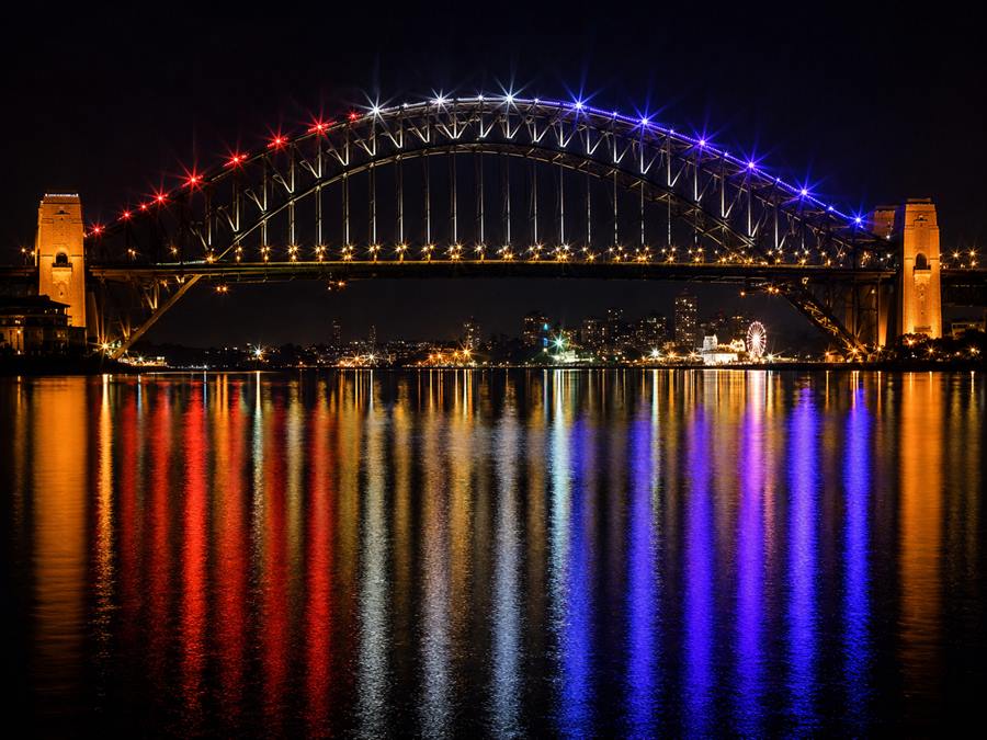 A steel arch bridge outlined in coloured LED lighting at night, reflected in the water below